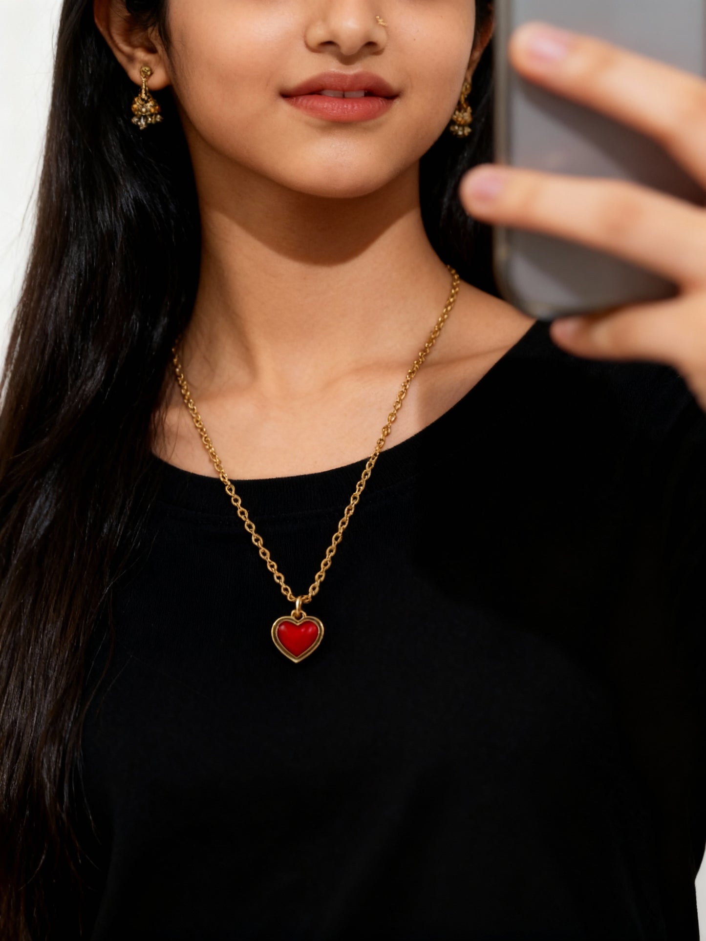 Woman wearing a gold necklace with a red heart pendant, taking a selfie.